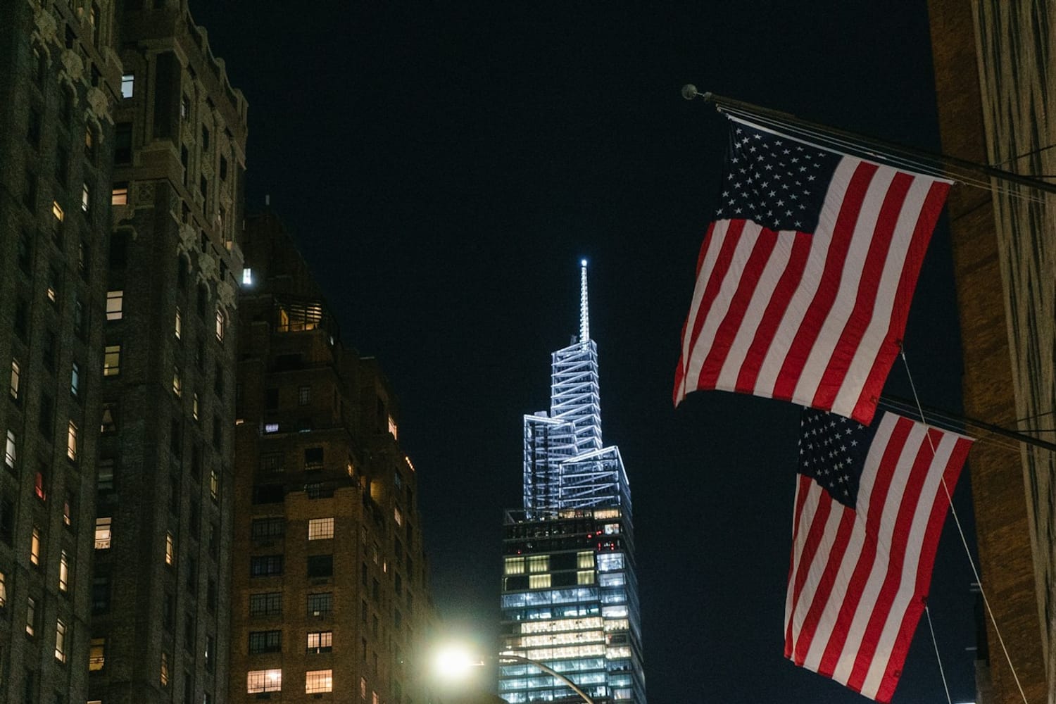 Two American flags on a building facade at night in New York, with an illuminated skyscraper behind