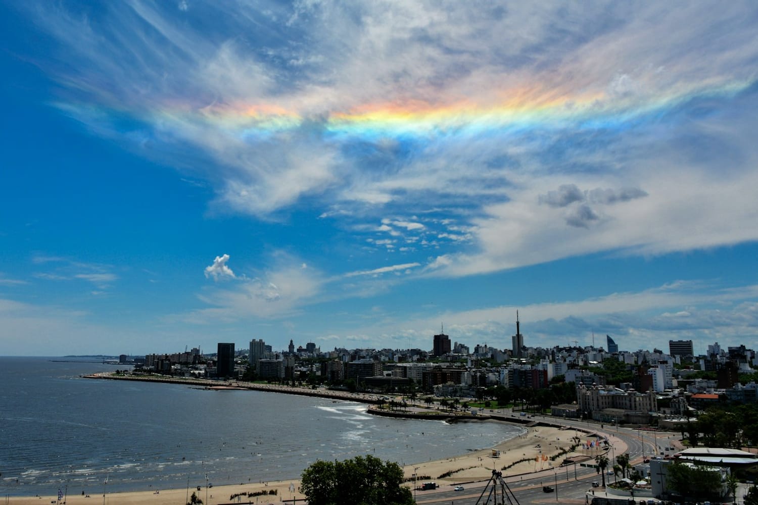 Montevideo's coastline and rambla under a blue sky with a rainbow-coloured cloud